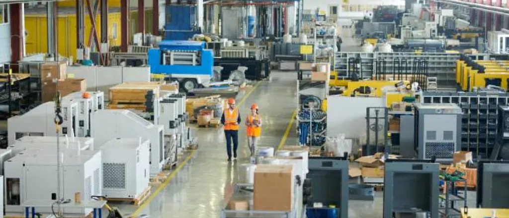 A large factory of machinery and heavy equipment with two workers in orange high-visibility vests and helmets walking through.