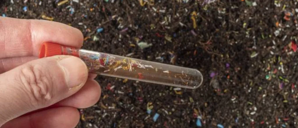 A hand holding a test tube containing a soil sample above a patch of dirt littered with colorful microplastic shards.