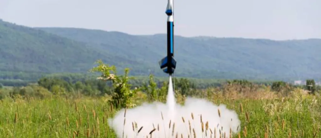 A blue and white colored model rocket launches in a cloud of smoke in the middle of field surrounded by rolling hills.