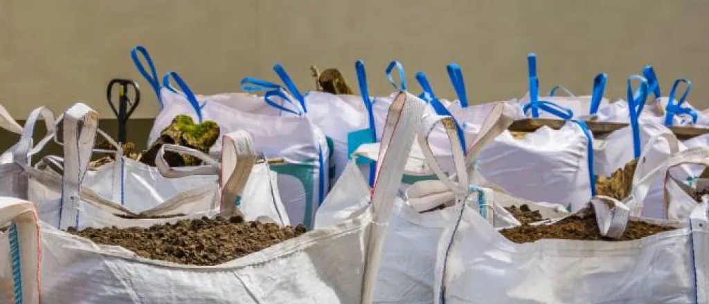 A group of white bags with blue stitching and handles gathered together and filled to the brim with potting soil.