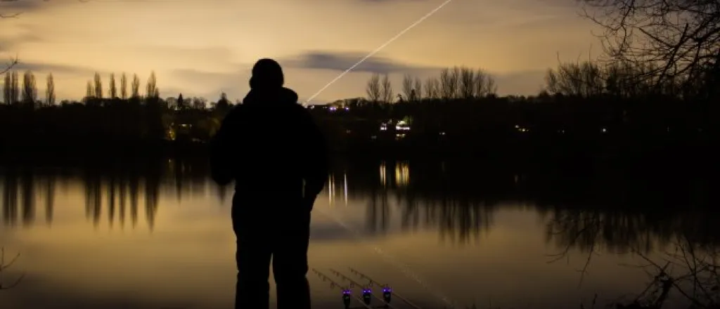 An angler standing at the edge of a lake just after sunset with three fishing rods resting on the ground next to him.