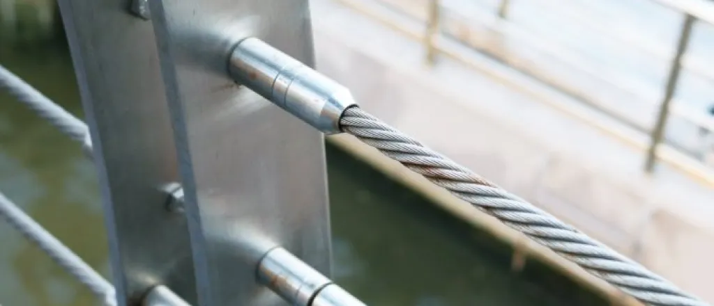 A close-up of a stainless steel threaded stud joint with wire rope cable railing over top of a body of water.