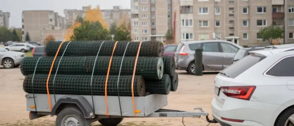 A parked white car with a trailer hitch and a utility trailer behind it carrying rolls of metal fencing strapped to the trailer.