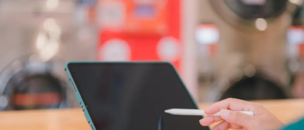 Someone using a stylus on their propped-up tablet at a laundromat. There are laundry machines blurred out in the background.