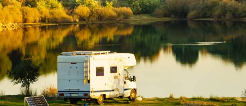 A recreational vehicle parked next to a pond or small lake with a solar panel behind it in a natural setting around sunset.