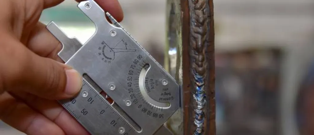 A technician uses a welding gauge to measure the dimensions of a metal part. The metallic joint lines are visible.