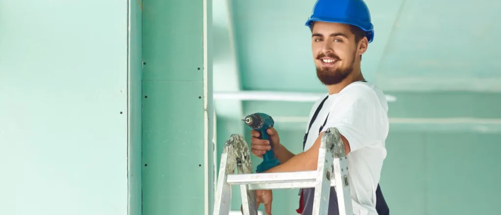 A male worker with a beard and mustache stands at the top of a ladder using a drill to install drywall sheets.