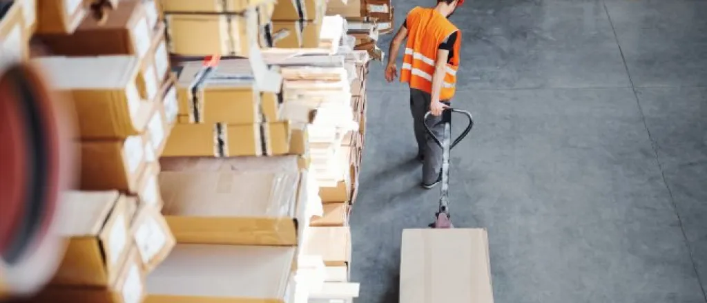 Aerial view of a warehouse worker pulling a cardboard box on a pallet truck next to shelving full of similar boxes and packages.