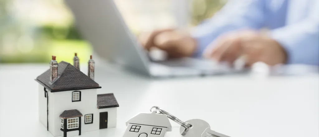 PROMO HIRES Miscellaneous - House Home Key Real Estate - iStock - BrianAJackson Miniature house and key on a keychain on a table. A person's hands and notebook computer are in the background.