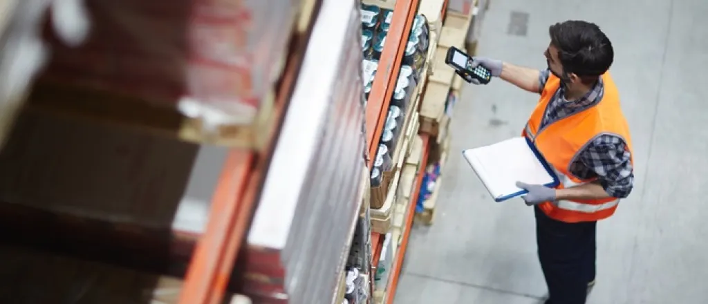Ways warehouses can improve their inventory systems A man in an orange vest is holding an electronic device up to a shelf filled with pallets with various items on them.