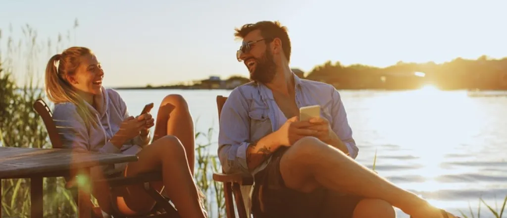 A young couple smiles and laughs as they sit on the side of a river. Both of them hold smartphones in their hands.