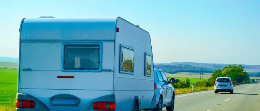 A white car towing a camper on a highway. There is an empty green field around the road and a clear blue sky overhead.