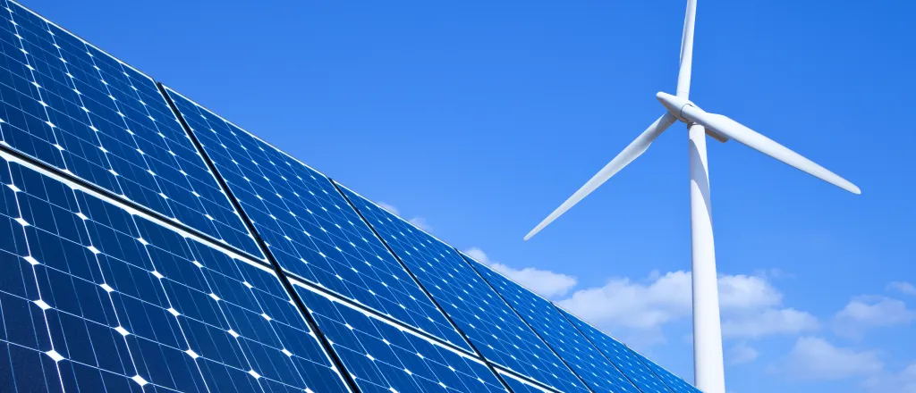 PROMO HIRES CROPPED Energy - Solar Panels Wind Turbine Renewable Climate - Eyematrix - iStock-453911335 Solar panels in the foreground, with a wind turbine in the background, against a blue sky with a few clouds.
