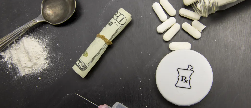 Overhead view of white powder next to a spoon, hypodermic syringe and needle, a rolled up United States 20 dollar bill with rubber band around it, and an open pill bottle with the contents partially spilled.