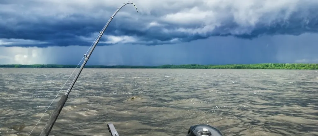 Angling 101: are fish most active before a storm? The stern of a boat on a lake before a storm hits. A fishing rod is cast into the choppy water below a dark, cloudy sky.