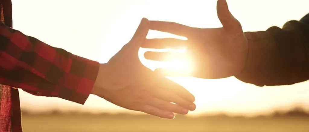 Two people shaking hands as the sun goes down behind them. They're standing on a piece of empty farmland.
