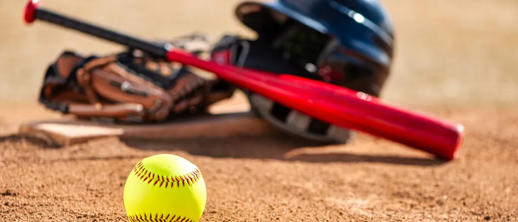 PROMO HIRES Sports - Softball School Bat Ball Glove Helmet - EHStock - iStock-2210228475 A softball sit in the foreground, with a helmet, bat, and glove sitting on a base slightly blurred in the background.