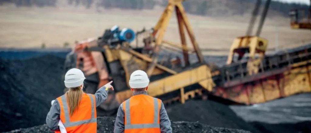 Two mining workers supervise a mechanical crane in a field of coal mounds. One of them gestures toward the machine.