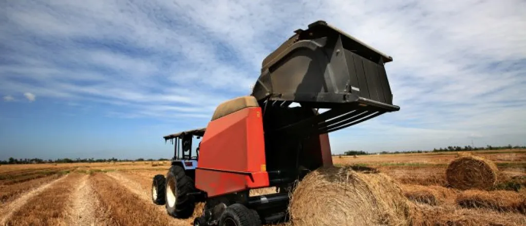 A rear view of a blue tractor towing a red round hay baler and dropping a rolled bale of hay onto a harvested field.