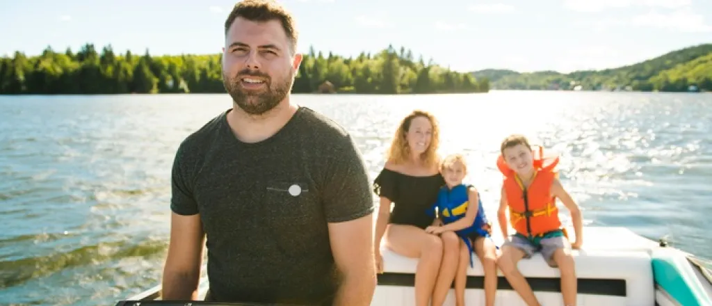 A man stands behind the steering wheel of a boat. A woman and two children sit behind the man at the back of the boat.