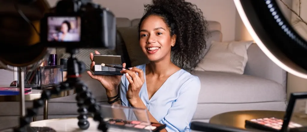 A young, Black influencer sits in front of a tripod and camera setup as she films a makeup eyeshadow palette.