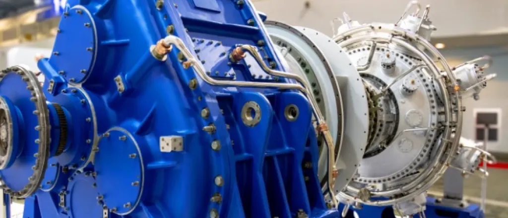A close-up of a large blue industrial gearbox connected to a turbine unit inside a factory with overhead lighting.