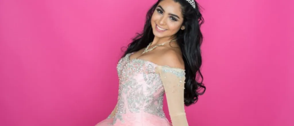 A girl with long, dark hair wearing a tiara and a formal pink dress poses against a bright pink background.