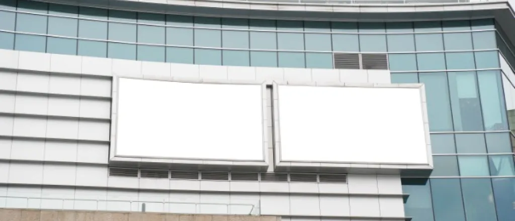 A low-angle view of two blank billboards hanging on the exterior of a large glass building on an overcast day.