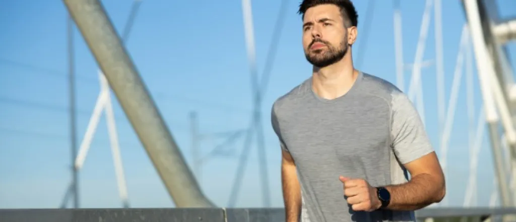 A man in a sweat stained shirt is jogging in an outdoor environment. Behind him is a suspension system for a bridge.