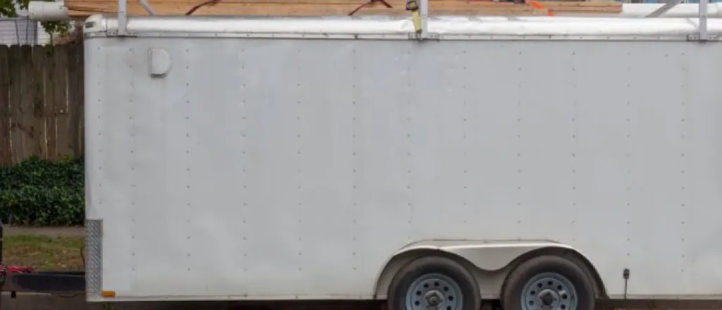 A white work trailer parked in a neighborhood on the curb. There is an orange ladder secured to the roof rack.
