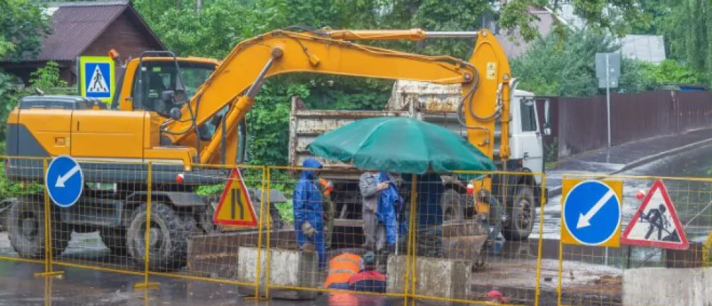 An excavator and dump truck parked behind a yellow fence with different signs. Crew members wear blue ponchos as it rains.