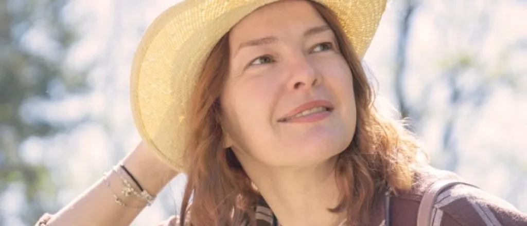 A woman wearing a straw hat and plaid shirt looking upward outdoors with trees and bright sunlight behind her.