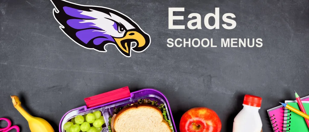 The Eads School Eagles mascot, chalked on a blackboard above a packed school lunch of a sandwich, grapes, carrots, an apple and a bottle of milk, with pencils and notebooks. The board reads “Eads School Menus”.