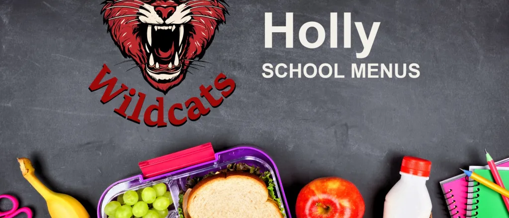 The Holly School Wildcats mascot, chalked on a blackboard above a packed school lunch of a sandwich, grapes, carrots, an apple and a bottle of milk, with pencils and notebooks. The board reads “Holly School Menus”.
