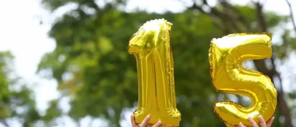 Two hands hold shiny gold number balloons forming 15 outdoors, with leafy green trees blurred in the background.