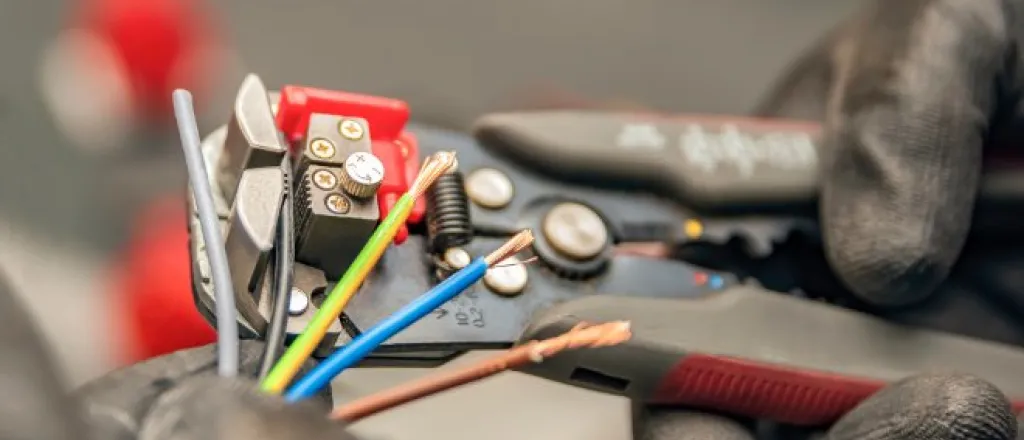 A close-up of a pair of hands with black gloves using a manual wire stripper to strip five colored electrical wires.