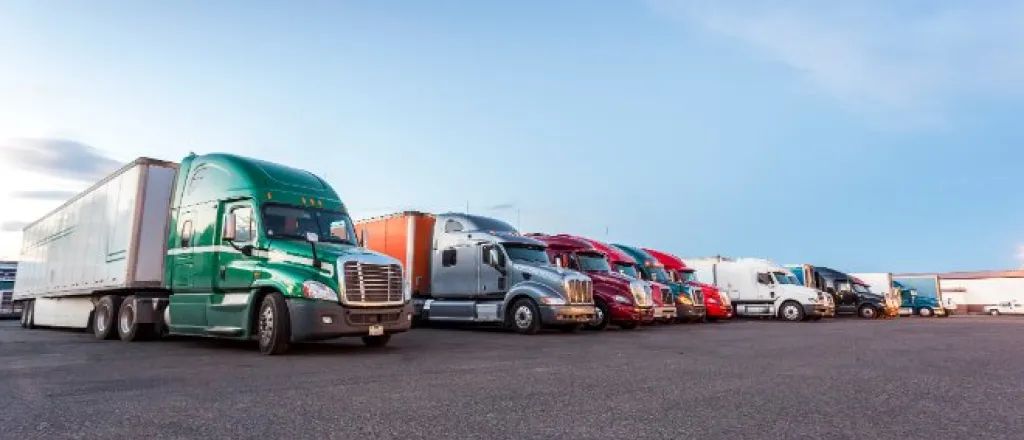 A row of colorful semi-trucks and trailers parked side by side in a large paved lot beneath a clear blue sky.