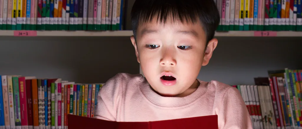 Child with a surprised expression holding an open book with glowing pages in front of books on shelves