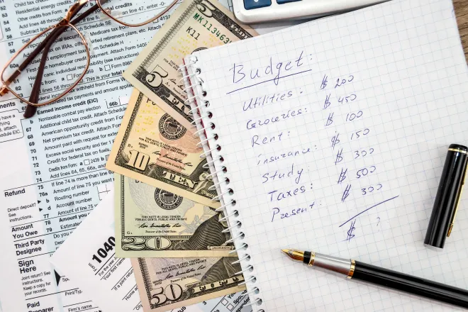 Notebook with a pen on a page showing a personal budget. Underneath are paper currency and tax forms. Eyeglasses are nearby