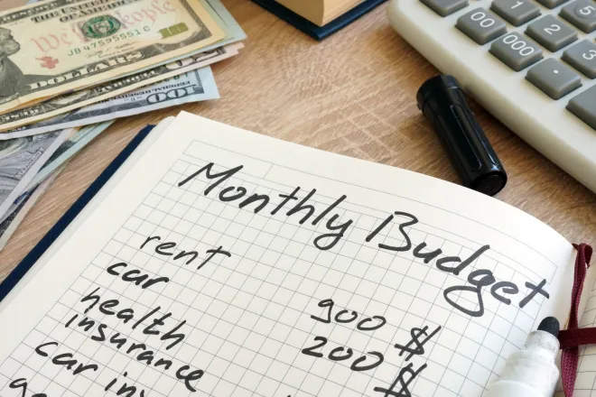 Notebook open to a page with hand-written "Monthly Budget" at the top. Cash and a calculator are nearby.