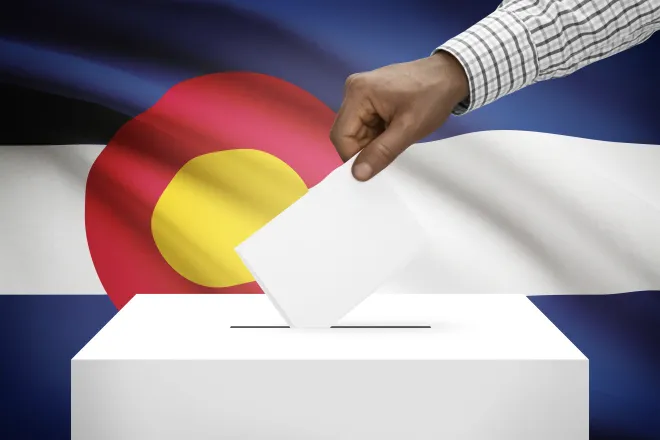 Hand inserting a piece of paper into a ballot box in front of the Colorado flag.