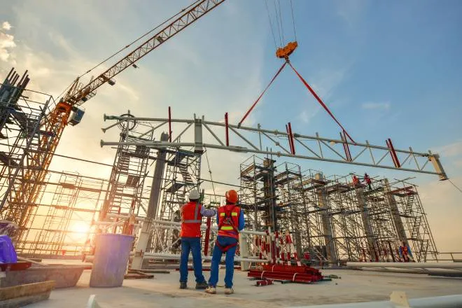 Two construction workers looking up as a steel truss is lifted by a large crane while the sun sets in the background.