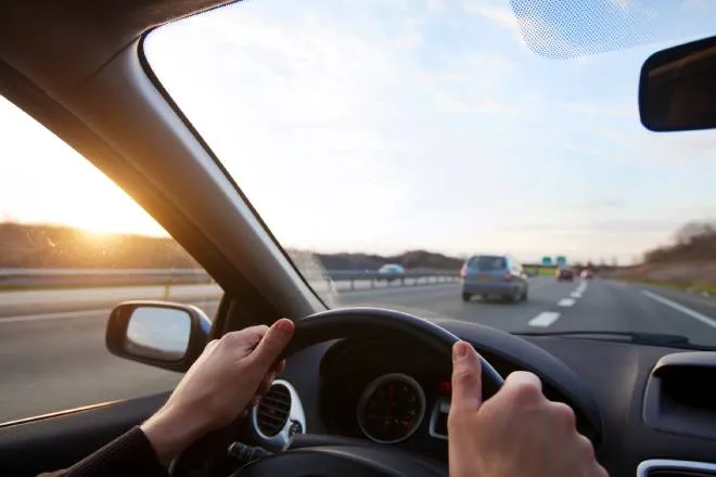 A person grips the steering wheel in their car as they drive on the highway with cars ahead of them and the sun setting.