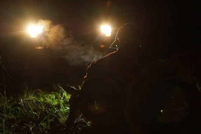 A hunter wearing a cap silhouetted in front of his truck's headlights as he carries the body of a deer toward his vehicle.