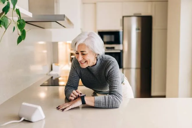 An older woman smiling as she leans over the kitchen counter to talk to her smart home assistant and adjust her smart watch.