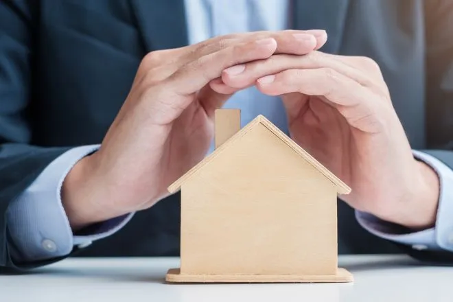 A man in a suit sitting at a table and covering the roof of a small wooden home model with both hands.