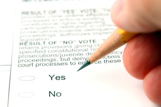 Fingers holding a pencil over an election ballot showing yes and no options