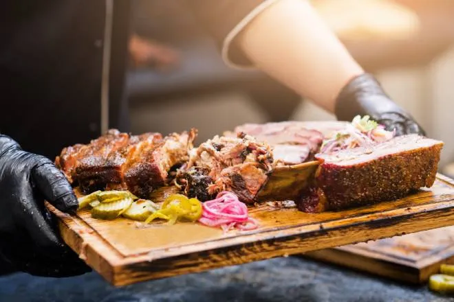 A man with gloved black hands holding a wooden tray of meat and vegetables in a professional restaurant setting.