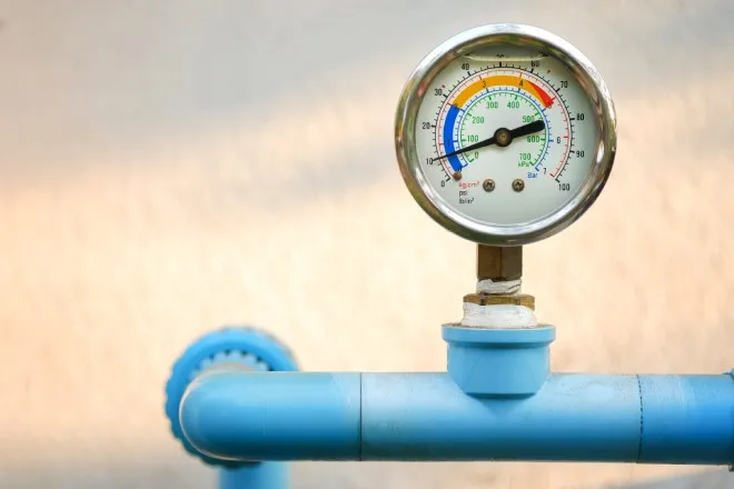 A pressure gauge with metal casing attached to a bright blue pipeline connected to an off-white wall.