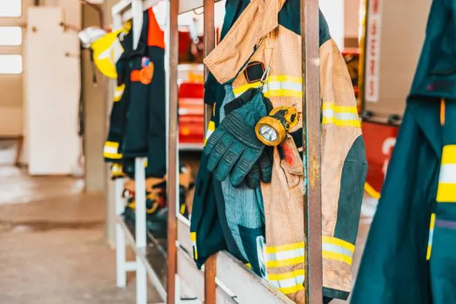 Fire-resistant clothing for firefighters, including jackets and gloves, hanging from a rusty metal rack.